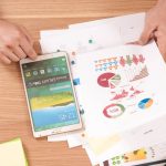 Person holding a white Samsung tablet and looking at a printout of charts