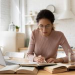 Concentrated young woman in eyewear reading carefully educational handbook, improving professional knowledge or preparing for university admission exams, writing notes in copybook, learning concept.