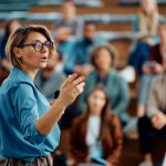 Mid adult businesswoman talking to group of seminar attendees in conference hall.