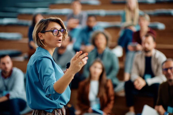 Mid adult businesswoman talking to group of seminar attendees in conference hall.