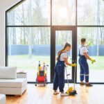 Young cleaners in blue uniform mopping wooden floor and cleaning big panoramic window