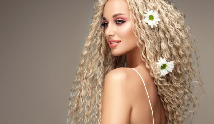 Portrait of young blonde haired beautiful model with flowers of chamomile in her long curly hairstyle. Soft smile on the attractive face.