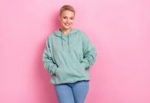 Photo promo poster of young satisfied charming blonde short hair girl wear put hands pockets khaki hoodie infuencer isolated on pink color background.