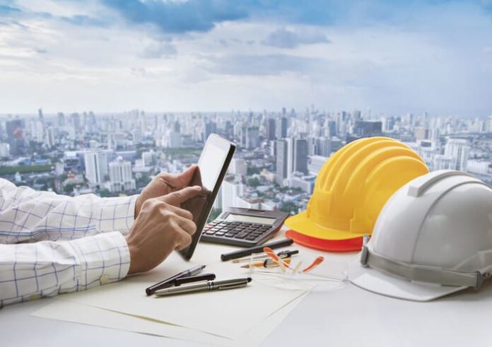 Hand of business man touching on computer tablet screen and safety helmet on working table against cities of high building