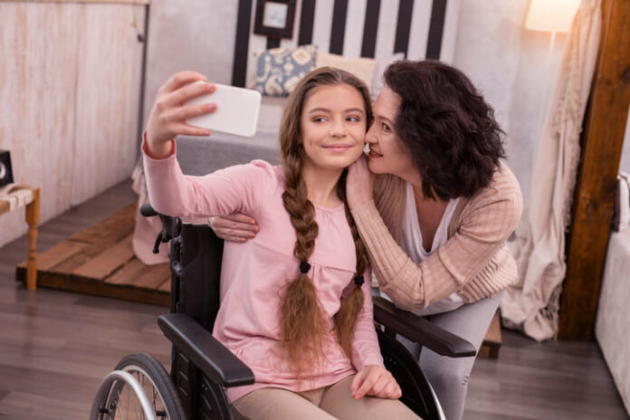 girl in wheelchair using phone to take a selfie while woman kissing her