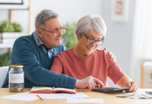 Elderly married couple sitting at the desk with a paper receipt in hands are calculating expenses, managing the budget.