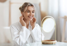 Cheerful beautiful blonde middle aged lady in silky bathrobe using hydrating face cream