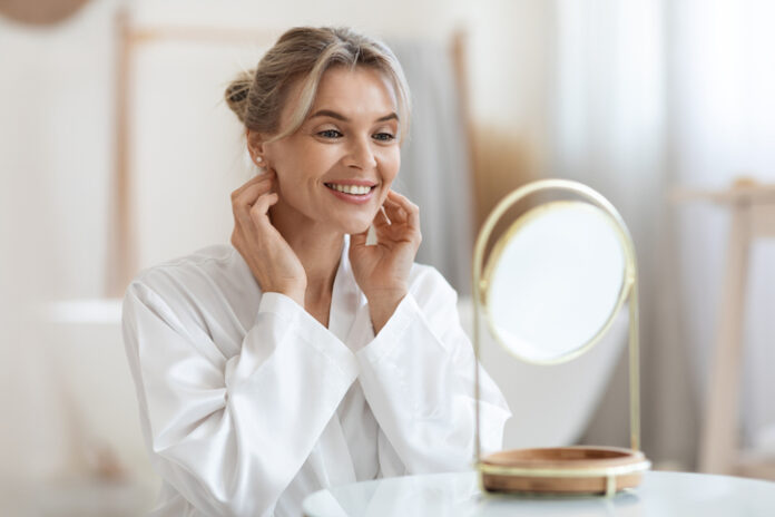 Cheerful beautiful blonde middle aged lady in silky bathrobe using hydrating face cream