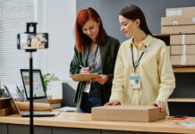 Young volunteer with packed parcel standing by desk next to her colleague with document while both looking through list of people in need