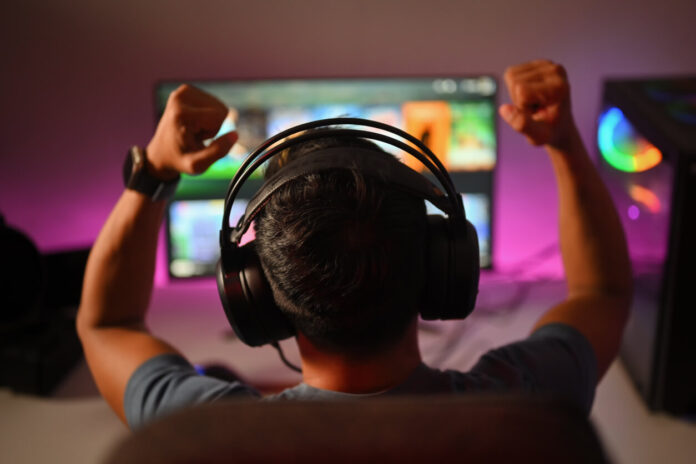 Back view of professional gamer wearing headphones celebrating victory while playing online video game at home