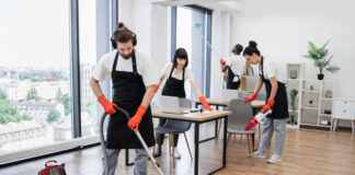 Team of young multicultural cleaners vacuums floor, wipes tables with gadgets, shelves, washes windows in spacious, bright, modern office. Professional cleaning company.