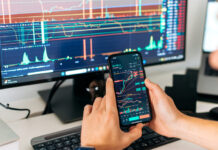 Male trader sits in home office in front of screens with graphs and financial charts, using smartphone