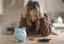 A woman sits at a kitchen table with her head in her hands, looking at a piggy bank and a calculator.