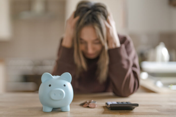 A woman sits at a kitchen table with her head in her hands, looking at a piggy bank and a calculator.