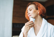 Woman In White Bathrobe With Red Hair Doing Fasial Massage With Microcurrent Facial Massager