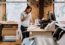 Portrait of charming woman in blue shirt looking at notes while standing near table with printer