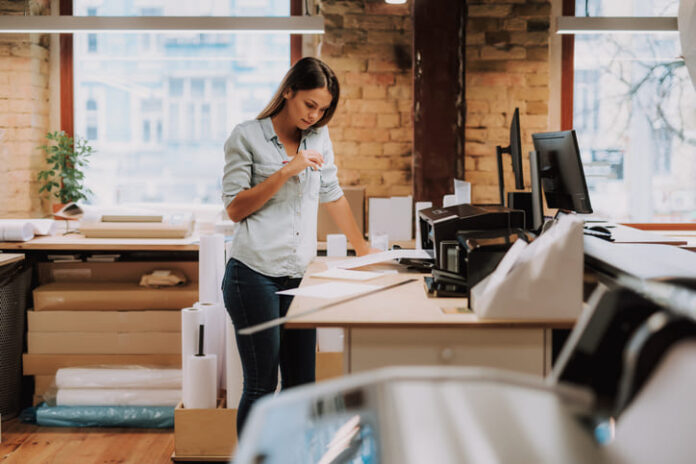 Portrait of charming woman in blue shirt looking at notes while standing near table with printer