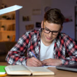 Student in glasses doing homework, writing essay, open book and laptop on table