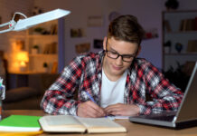 Student in glasses doing homework, writing essay, open book and laptop on table