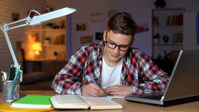 Student in glasses doing homework, writing essay, open book and laptop on table