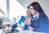 Businesswomen working together finger pointing at laptop on desk, smiling portraits. Glowing padlock icon with circuit hud. Cybersecurity and data protection.