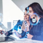 Businesswomen working together finger pointing at laptop on desk, smiling portraits. Glowing padlock icon with circuit hud. Cybersecurity and data protection.