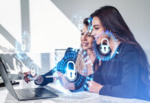 Businesswomen working together finger pointing at laptop on desk, smiling portraits. Glowing padlock icon with circuit hud. Cybersecurity and data protection.