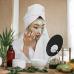 Woman looking at mirror sitting at table with ingredients for homemade cosmetics applying green moisturizing mask on her face