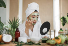 Woman looking at mirror sitting at table with ingredients for homemade cosmetics applying green moisturizing mask on her face