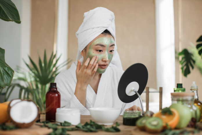 Woman looking at mirror sitting at table with ingredients for homemade cosmetics applying green moisturizing mask on her face