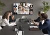 Employees speaking online to diverse freelancers on wall display and office workers at meeting table