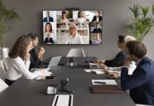 Employees speaking online to diverse freelancers on wall display and office workers at meeting table