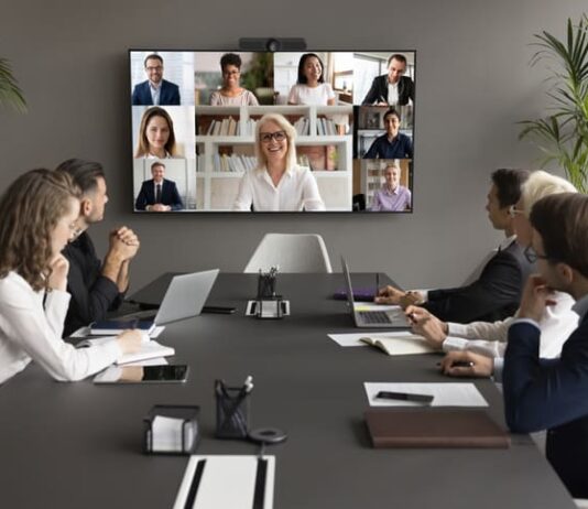 Employees speaking online to diverse freelancers on wall display and office workers at meeting table