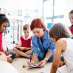 A group of small school kids with teacher sitting on the floor in class, learning