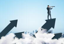 Businessman standing on abstract arrow and using telescope to look into the distance on city background.