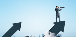 Businessman standing on abstract arrow and using telescope to look into the distance on city background.