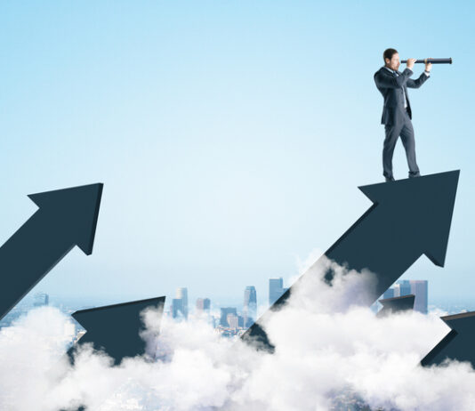 Businessman standing on abstract arrow and using telescope to look into the distance on city background.