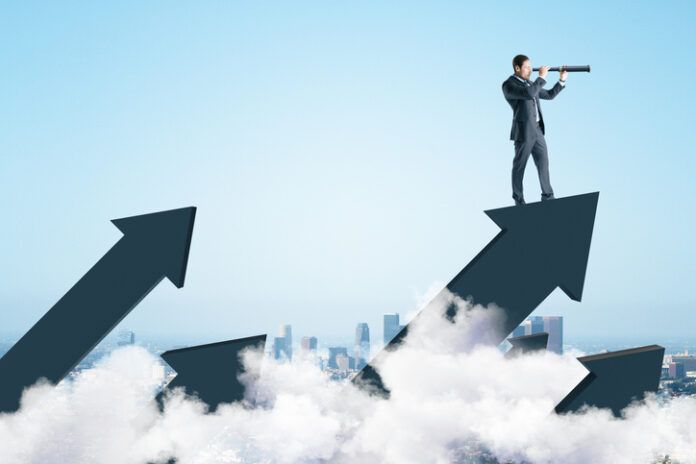 Businessman standing on abstract arrow and using telescope to look into the distance on city background.