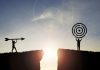 Silhouette men throwing arrow and holding dartboard on mountain with blue sky and sunlight for achievement objectives target