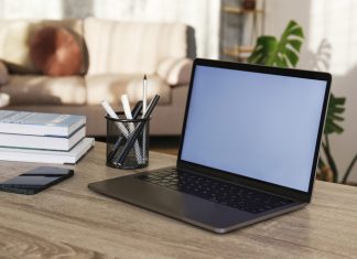 Blank screen laptop, cell phone and a cup on wooden table. Cozy lofty office with house plants full of natural sunlight