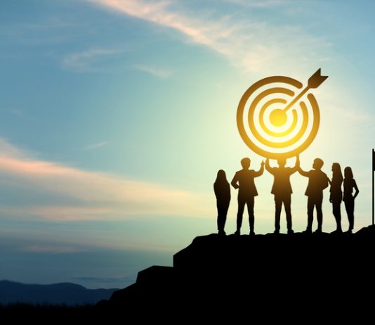 Silhouettes of group businessmen holding target boards with flags planted on a mountain.