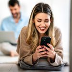 Happy smiling young woman holding smartphone on floor at home using online entertainment mobile app