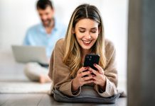 Happy smiling young woman holding smartphone on floor at home using online entertainment mobile app