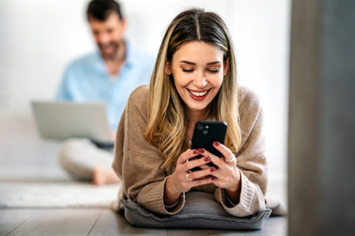 Happy smiling young woman holding smartphone on floor at home using online entertainment mobile app