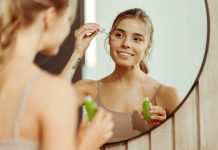 Young woman holding green bottle applying serum to face, looking in mirror, standing in bathroom